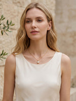 A blonde model wearing a white sleeveless top, standing against a textured wall with greenery, modeling the gold oval pendant necklace.